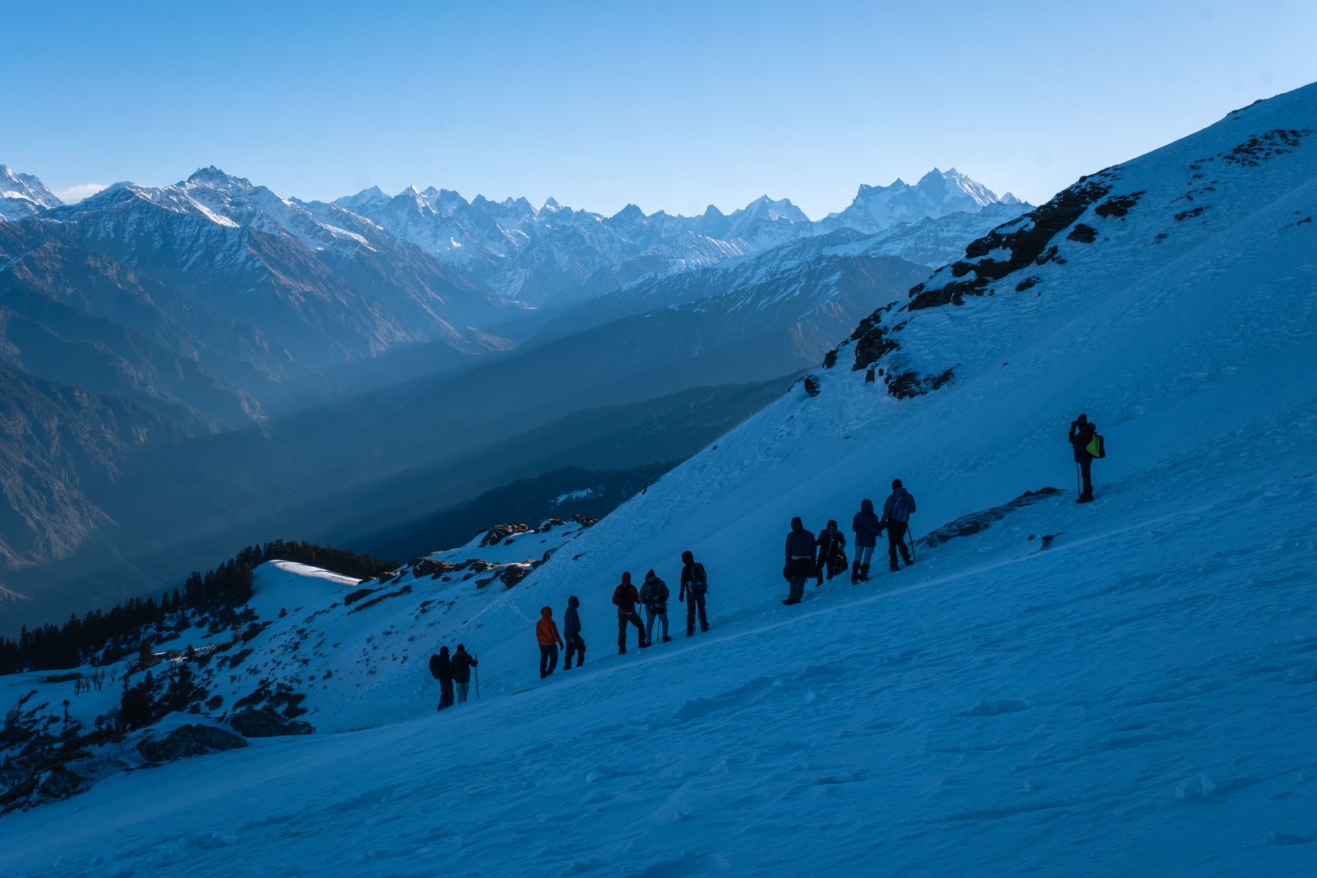 A line of trekkers crossing a snowy mountain ridge at dusk, with a distant mountain range beyond the valley