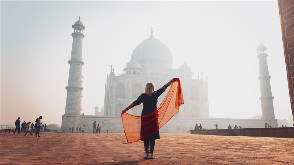 A woman in an orange scarf walking in front of the Taj Mahal in hazy morning light