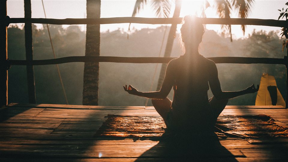 A person meditating in silhouette on a wooden deck, backlit at sunset